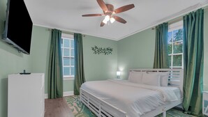 Un dormitorio con cama, ventilador de techo, televisión y ventana con cortinas.