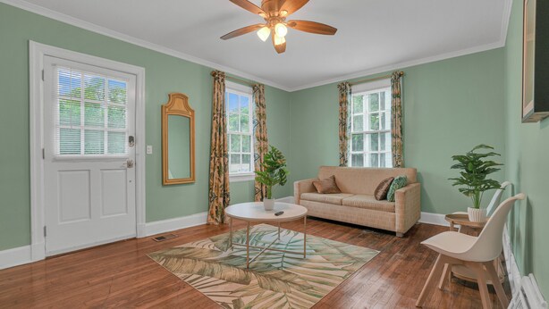 Una sala de estar con un sofá color beige, una mesa de centro y un ventilador de techo. Hay dos ventanas con cortinas, una puerta y un espejo en la pared.