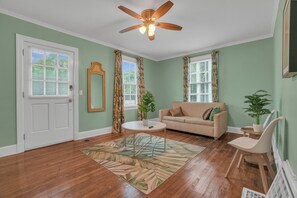 A living room with a beige sofa, a coffee table, and a ceiling fan. There are two windows with curtains, a door, and a mirror on the wall. - Cozy Green Haven, 1BR Flat Minutes from Downtown (Montgomery)