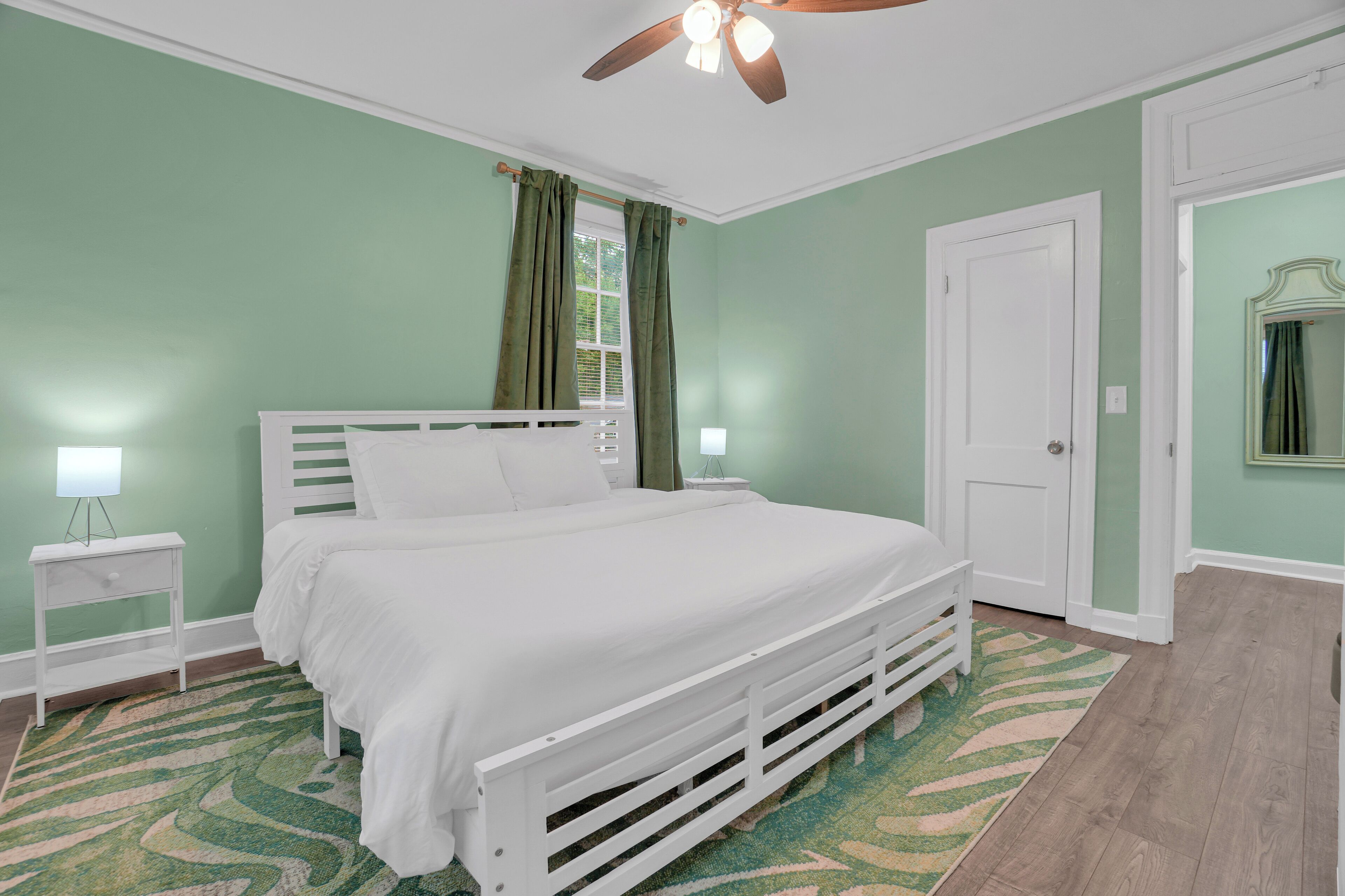 Une chambre à coucher comprenant un lit, des tables de chevet, un ventilateur de plafond, une porte et un miroir.
