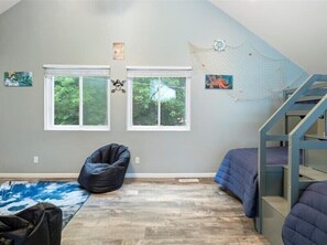 A room with two windows, a black bean bag chair, a blue bed, and a wooden floor.