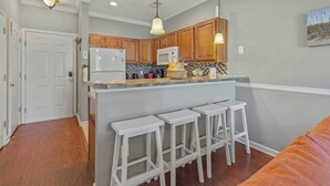 A kitchen with wooden cabinets, a white refrigerator, a microwave, and a granite countertop. There are three white stools at the island. - River Oaks Unit 63C -Near Golf Areas! (Myrtle Beach)