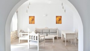 Una habitación con techo abovedado blanco, un sofá blanco, una mesa blanca y una silla blanca. Hay dos cuadros en la pared y una ventana con cortinas.