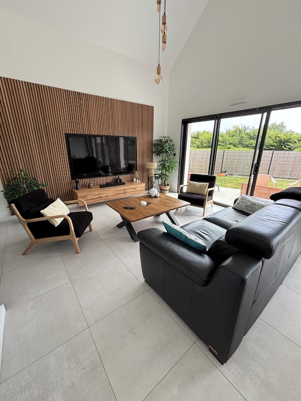 A modern living room with a flat-screen TV, a wooden accent wall, a large black sofa, a coffee table, and a view of the outdoors.