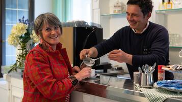 Una persona serve caffè all'altra al bancone, con una macchina da caffè, un vaso di fiori e una scaffalatura con bottiglie sullo sfondo.