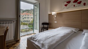 A hotel room with a bed, a chair, a radiator, a window with a view of buildings, and a wall-mounted lamp. - Hotel Vecchie Mura  (Finale Ligure)