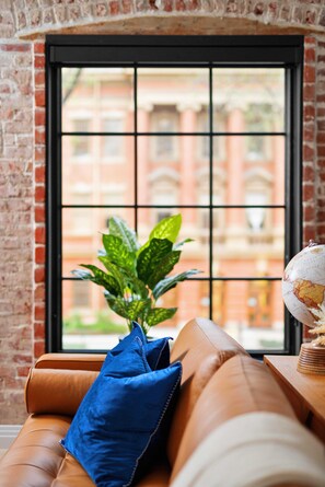 A leather sofa with a blue pillow and a potted plant in front of a window with a brick wall and a view of buildings. - Alex Pangburn Suite 212 (Denver)