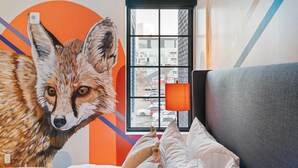 Een kamer met een grote muurschildering van een vos, een raam met uitzicht op gebouwen, en een grijze bank met een wit kussen.