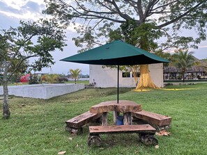 A picnic table with a green umbrella in a grassy area with trees and a building in the background. - Hotel Zensi 420 (Lejanías)