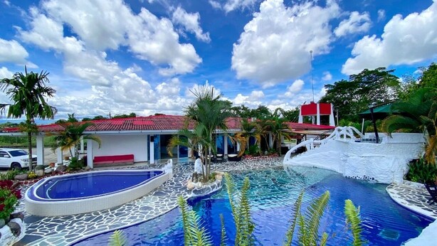 A resort pool area with a clear blue sky, palm trees, and a building with a red roof. - Hotel Zensi 420 (Lejanías)