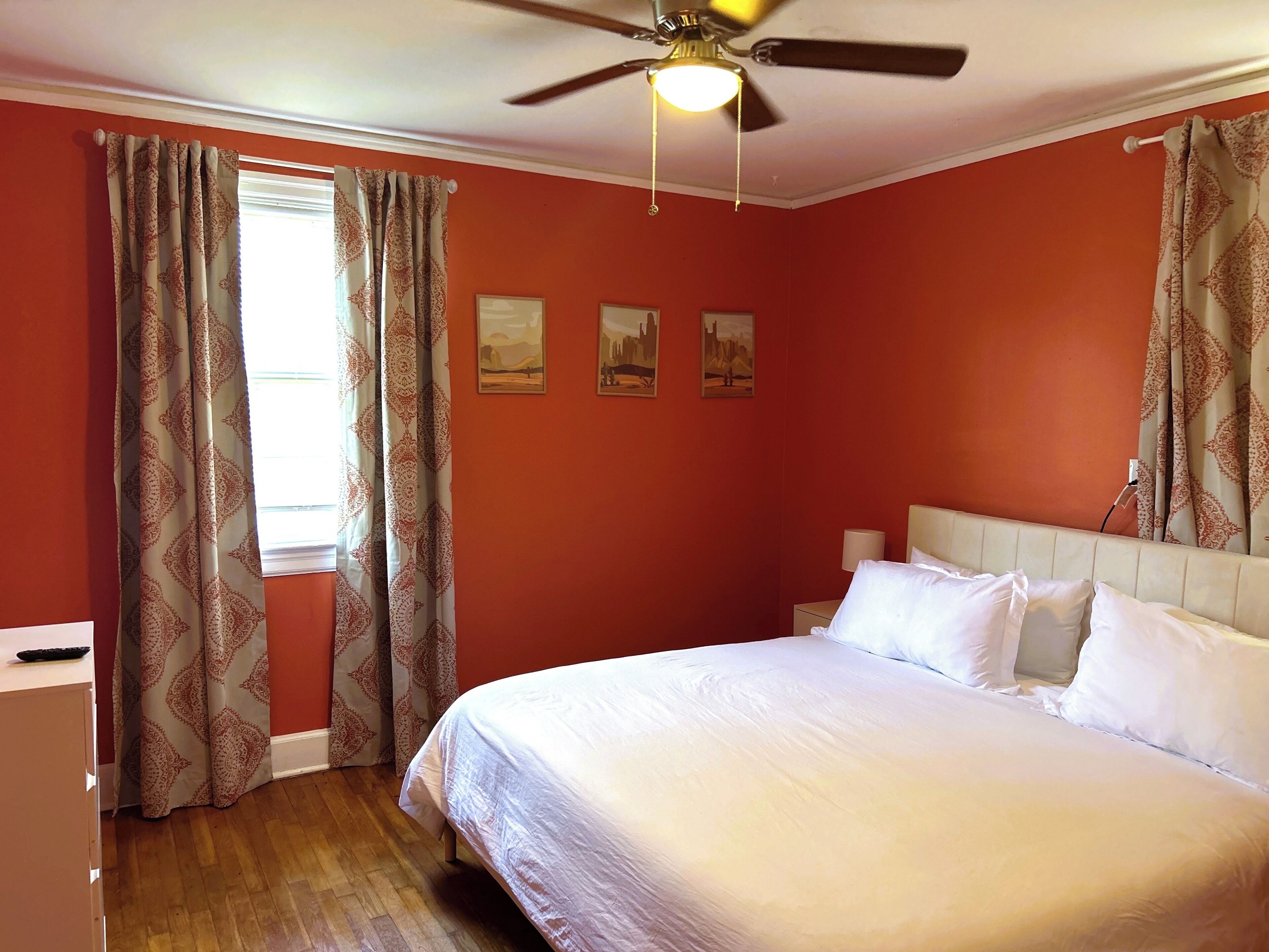 A bedroom with a bed, curtains, a ceiling fan, and wall art.
