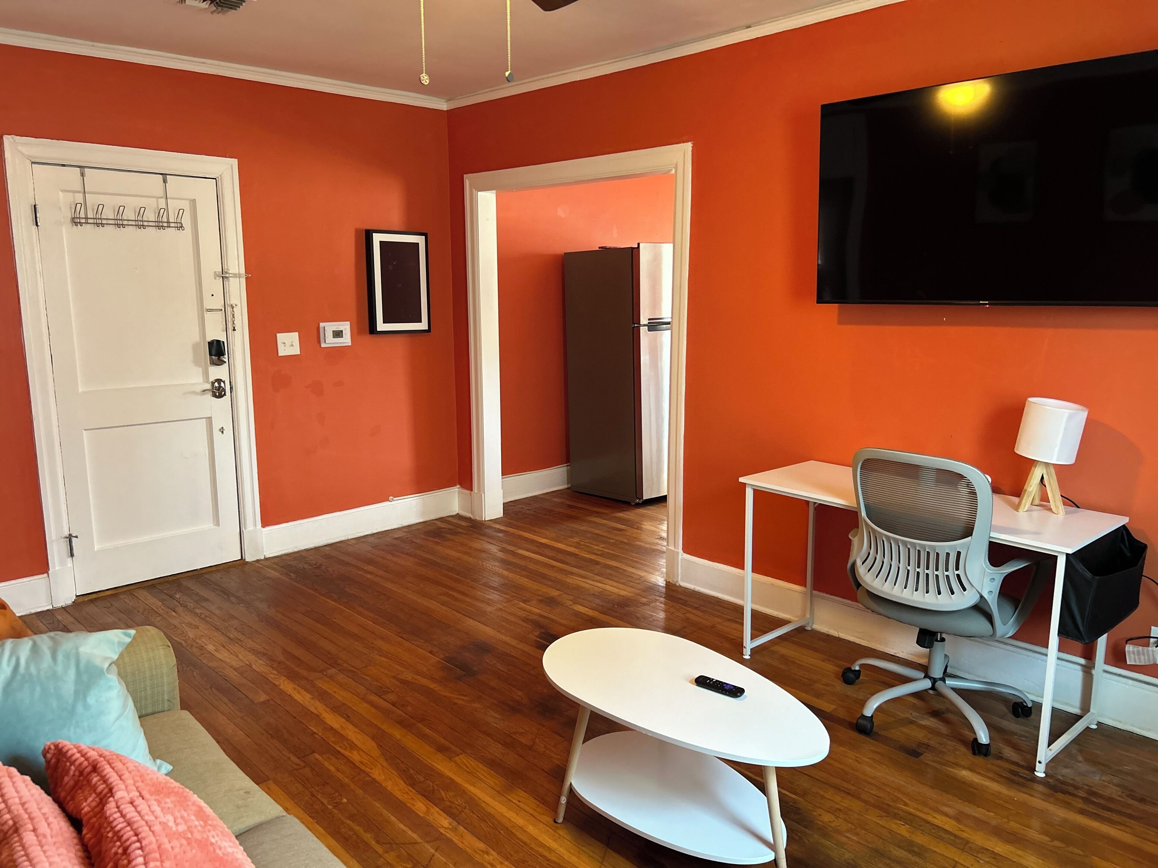 A room with a wooden floor, a white door, a white desk with a chair, a TV mounted on the wall, and a framed picture on the wall.