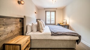 A bedroom with a wooden headboard, a bed with white and gray bedding, a bedside table, and a window with a view of the outdoors. - Kotibe- Lovely apartment in Chamonix (Chamonix-Mont-Blanc)