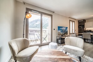 A modern living room with a large sliding glass door leading to a balcony, a wooden coffee table, two armchairs, and a painting on the wall.
