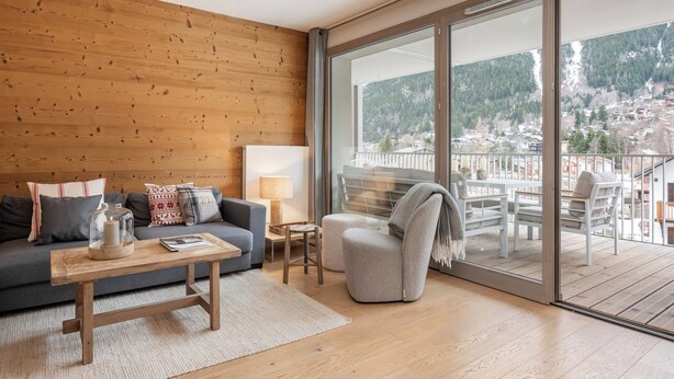 Een moderne woonkamer met een houten wand, een grijze bank, een houten salontafel en uitzicht op een balkon met tuinmeubilair.