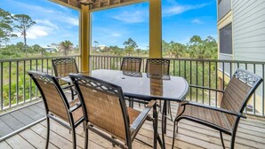 A covered outdoor patio with a round table and six chairs, surrounded by a wooden deck and metal railing, overlooking a residential area.
