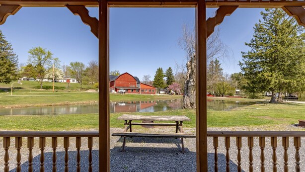 Una pérgola de madera con suelo de grava, con vista a un estanque y un granero rojo.