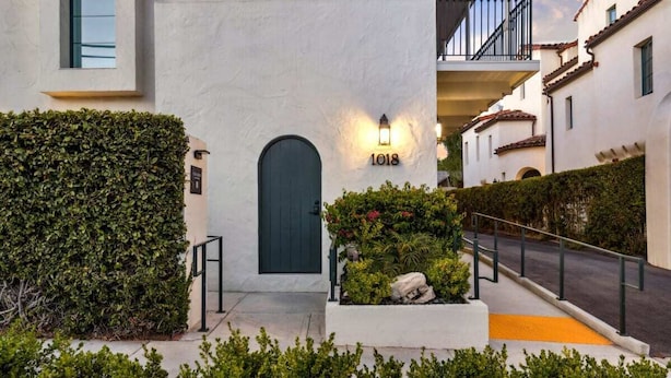 A building with a dark door, a number '1018' sign, a light fixture, and a small garden area with plants and rocks.
