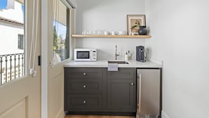 A compact kitchen with dark cabinets, a microwave, a sink, and a small refrigerator. There is a window with a view of the outside, and a shelf above the sink holding dishes and a framed picture.