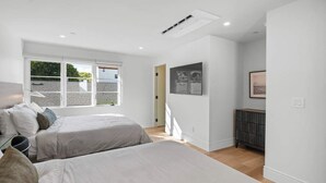 A modern bedroom with two beds, a large window, and a small cabinet.
