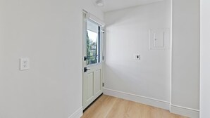A narrow hallway with white walls and a wooden floor. There is a door with a window on the left, a light switch on the right, and a ceiling vent.