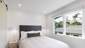 Een slaapkamer met een bed, bedlampen, een raam met uitzicht op gebouwen en bomen, en een witte muur.