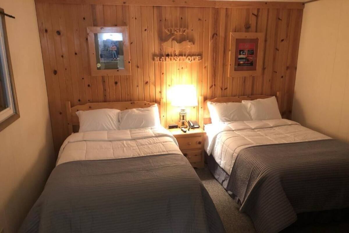 A room with two beds, wooden panel walls, a framed picture, and a lamp on the nightstand.