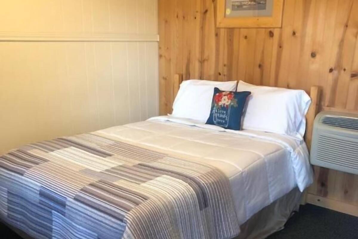 A single bed with a striped bedspread, white pillows, and a small decorative pillow. A framed picture hangs on the wooden wall above the bed.