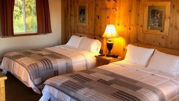 A room with two beds, wooden walls, a window with curtains, and a lamp on a nightstand. - Holiday Motel (Manistique)