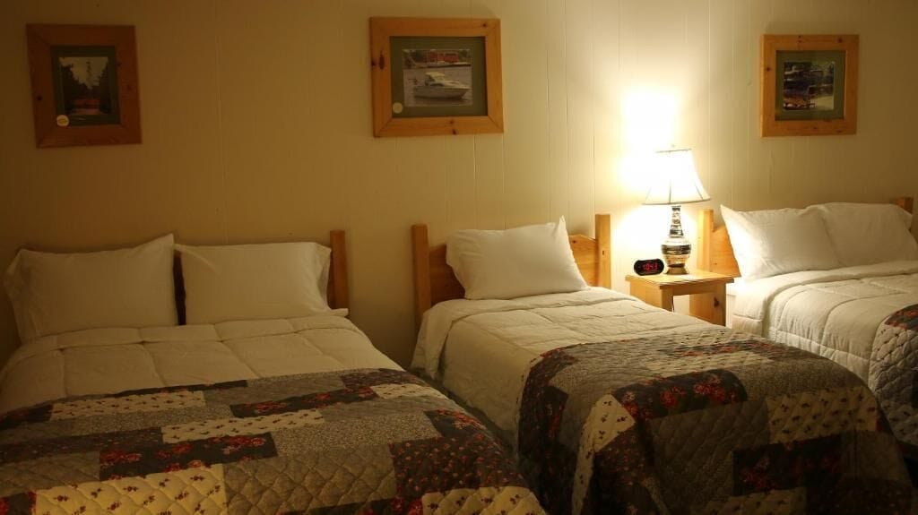 A hotel room with three single beds, each with a quilted bedspread and a wooden headboard. There is a nightstand with a lamp between the beds, and framed pictures on the wall above each bed.