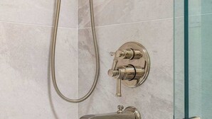 Ducha con cabezal de ducha de mano y grifo empotrado en la pared, ambos con acabado metálico, frente a una pared azulejada.