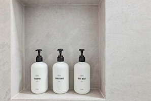 Drie witte flessen van Public Goods shampoo, conditioner en douchegel op een witte plank.