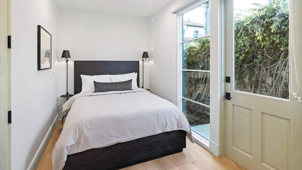 Ein Schlafzimmer mit einem Bett, zwei Nachttischlampen, einem Fenster mit Blick auf Grünflächen und einer Tür.