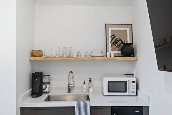 Une cuisine compacte dotée d’un plan de travail blanc, d’un évier en acier inoxydable, d’un four à micro-ondes, d’une machine à café et d’une étagère où sont rangés des verres et une photo encadrée.