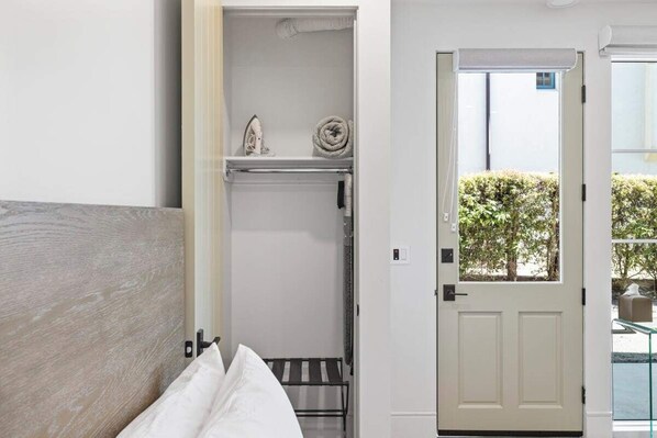 A room with a bed, a white door, a small shelf with an iron and a rolled towel, and a view of greenery outside.