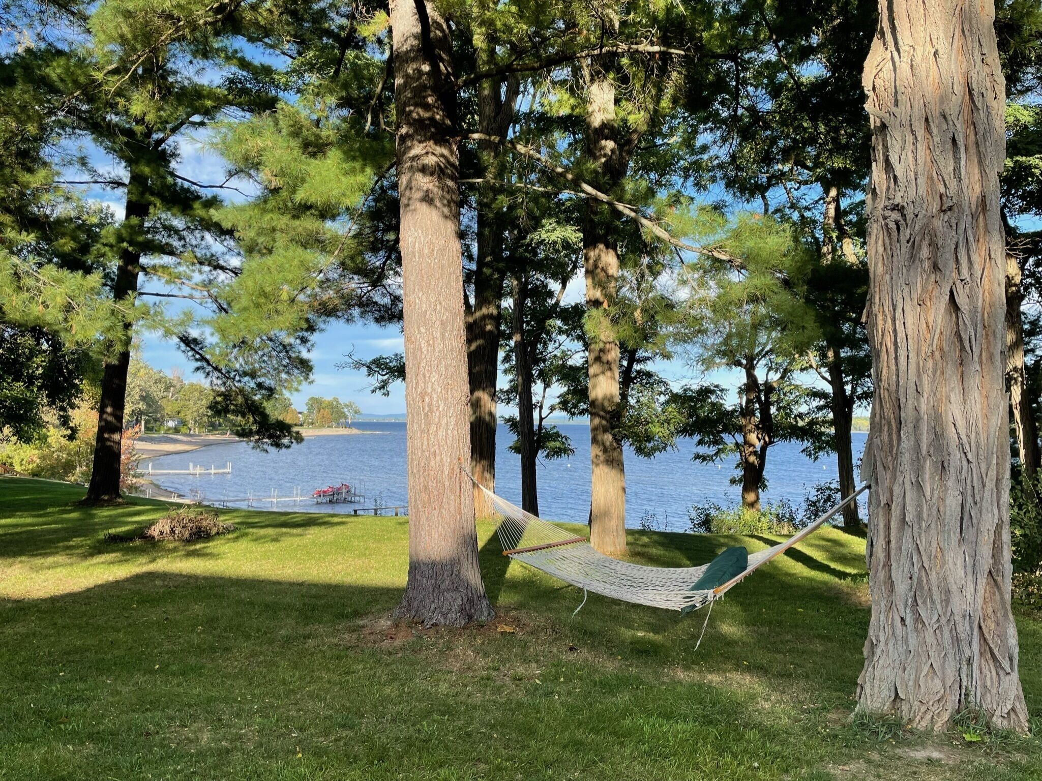 Un hamac tendu entre deux arbres, près d’un lac, avec une jetée et un bateau en arrière-plan.
