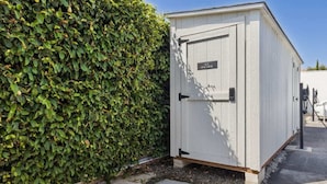 Un abri de jardin blanc, utilisé pour ranger des outils, avec une porte sur laquelle on peut lire « MACHINE À AIR », situé à côté d’une haie verte et touffue.