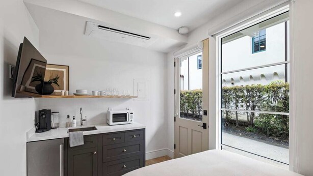 Un dormitorio moderno con una cama grande, un armario empotrado, una pequeña cocina americana y una puerta corrediza de vidrio que da a un jardín.