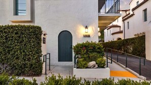 Un edificio con una puerta oscura, un cartel con el número 1018, un accesorio de iluminación y un pequeño jardín con plantas y rocas.