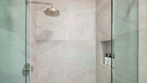 Ducha con paredes de mármol y mampara de vidrio, incluye un cabezal de ducha de mano y un pequeño estante con artículos de tocador.