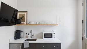 Una cocina moderna con encimera blanca, una cafetera negra, un microondas blanco y un estante sobre el fregadero con platos y vasos.