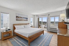Quarto moderno com cama de madeira, mesa de cabeceira, abajur, pintura na parede e vista pelas portas da varanda.