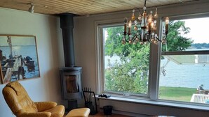 A cozy living room with a wooden ceiling, a large window with a view of a lake, a yellow armchair, a round table, and a patterned rug.