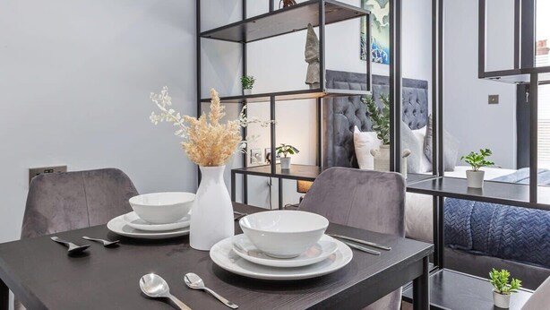 A modern dining area with a dark wooden table set for two, white bowls, and silverware. A white vase with dried flowers is on the table. Behind the table, there is a grey upholstered bench and a black metal shelving unit with decorative items. - Serviced Modern Luxury Flat near Central London (Greater London)