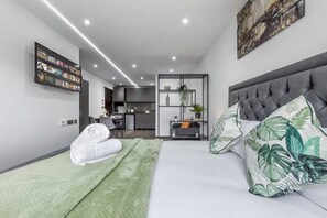 A modern hotel room with a large bed, a TV mounted on the wall, a small kitchenette, and a view of the cityscape.