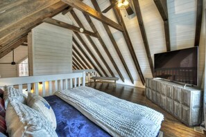 A bedroom with a sloped ceiling, a bed with a patterned bedspread, a television on a wooden stand, and a bench. - Picture Perfect | Hot Tub + Fireplace + Mtn Views (Blue Ridge)