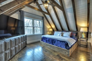 Ein Schlafzimmer mit schrägverdachter Decke, einem Bett, einem Fernseher und einem Fenster.
