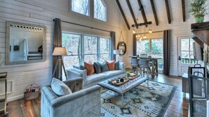 A spacious living room with a large sofa, coffee table, and a patterned rug. There are multiple windows providing natural light and a view of the outdoors. - Picture Perfect | $179 Off Your Stay! (Blue Ridge)