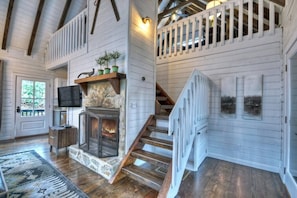 A spacious living area with a stone fireplace, wooden staircase, and a white railing.