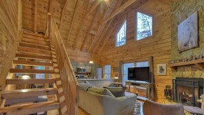 Ein Holzhütten-Innenraum mit Treppe, Sofa, Fernseher, Kamin und Küchenbereich.